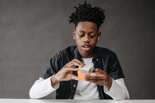 Young black teenager solving a Rubik's Cube in a studio setting on a gray background.